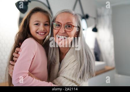 Ältere Großmutter und Enkelin stehen drinnen im Badezimmer, umarmen und betrachten die Kamera. Stockfoto