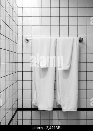 Zwei saubere weiße Handtücher gefaltet hängen an einem Metall Kleiderbügel Bar weißen Gittermuster Fliesen Wand an der Ecke des Badezimmers Hintergrund, minimalistischen Stil, vertica Stockfoto
