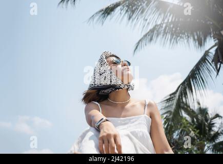 Schönes glückliches asiatisches Frauenportrait in lässigem weißen Hemd mit Sonnenbrille und Haarschal und Blick auf die Sonne am Strand unter dem Coconu Stockfoto