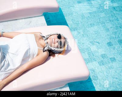 Attraktive glückliches Lächeln Asiatische Frau Porträt in lässigen weißen Hemd tragen Sonnenbrille und Haar Schal auf rosa Sonnenliege in der Nähe des Pools lehnen, genießen Sie die Stockfoto