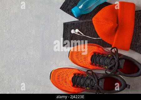 Orangefarbene Sneaker, ein orangefarbener Strickhut, warme Leggings für den Sport, eine Flasche Wasser und weiße Kopfhörer auf einem hellen Betontisch. Winter Running-Geräte Stockfoto