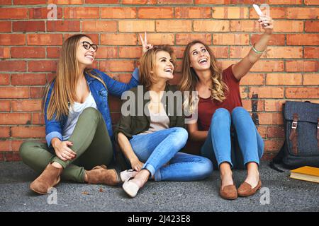 Erinnerungen an den Campus schaffen. Aufnahme von drei lächelnden Universitätsstudenten, die zusammen ein Selfie machen, während sie draußen auf dem Campus sitzen. Stockfoto