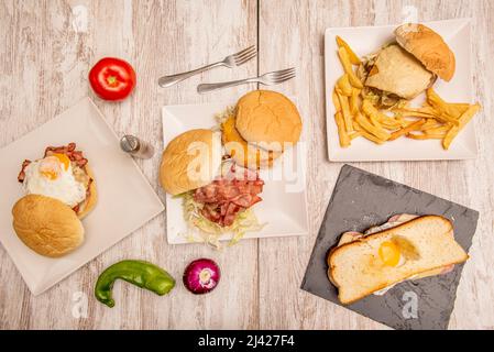 Fastfood-Gerichte mit verschiedenen Burgern, hausgemachten pommes frites, Sandwich mit Spiegelei und Tomaten und Paprika auf dem eingelegten Holztisch Stockfoto