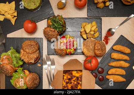 Fastfood-Gerichte mit verschiedenen Hamburgern, hausgemachten Pommes frites, Hähnchenfingern, Kartoffeln mit Frischkäse und Schinkentacos, Nachos mit Guacamole und frisch Stockfoto