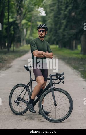 Radprofi in Sportbekleidung, Helm und Brille, der mit seinem schwarzen Fahrrad im grünen Park steht, zur Seite schaut und die Arme gekreuzt hält. Konzept von Stockfoto