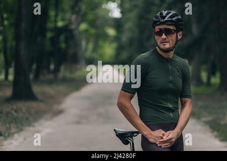 Porträt eines gutaussehenden männlichen Athleten mit Schutzhelm und verspiegelter Brille, der mit schwarzem Fahrrad im Freien posiert. Morgens Radfahren im grünen Park. Kopieren Sie den SPAC Stockfoto