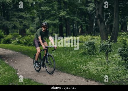 Starker junger Mann, der am Morgen auf dem schwarzen Fahrrad im grünen Stadtpark fährt. Aktiver kaukasischer Mann in Schutzhelm und Spiegelbrille, der alleine auf Fr. Stockfoto