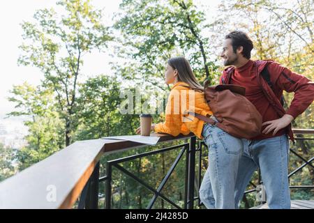 Seitenansicht eines lächelnden Reisenden, der seine Freundin mit Rucksack, Kaffee zum mitnehmen und eine Karte im Freien sah Stockfoto