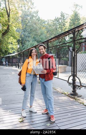 Positive Touristen mit Fernglas, Karte und Rucksäcken, die auf der städtischen Straße auf die Kamera schauen Stockfoto