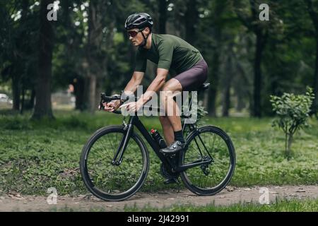 Seitenansicht des männlichen Athleten in Sportkleidung, Schutzhelm und verspiegelter Brille beim Fahrradfahren im Sommerpark. Konzept von Ausdauer, Selbstdisziplin und m Stockfoto