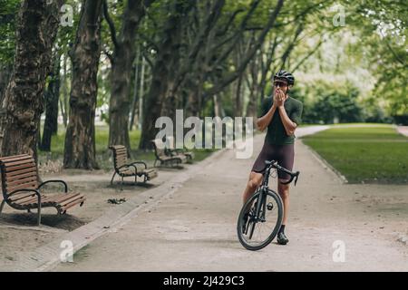 Müde Sportler in Sportbekleidung, Helm und Brille stehen mit Fahrrad im grünen Sommerpark. Junger muskulöser Kerl, der morgens Zeit für Outdoor-Aktivitäten verbringt Stockfoto
