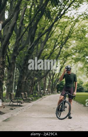 Junger Radfahrer mit muskulösen Beinen posiert im grünen Stadtpark mit einem professionellen schwarzen Fahrrad. Aktiver Mann in Helm und Sportkleidung Training regelmäßig aus Stockfoto