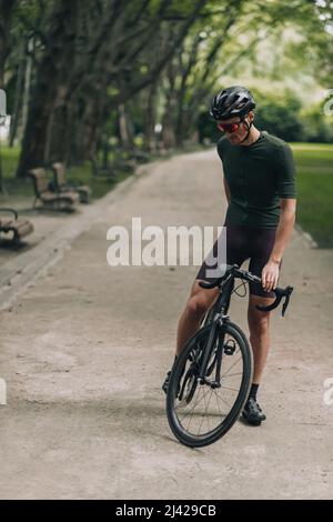 Starker kaukasischer Mann in aktiver Kleidung und Schutzhelm sitzt auf schwarzem Fahrrad im grünen Sommerpark. Konzept von Menschen, Sport und Erholung. Stockfoto