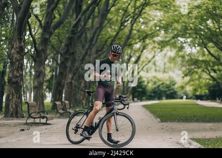 Porträt eines müden Radfahrers, der auf einem schwarzen Fahrrad sitzt und eine Sportflasche mit frischem Wasser hält. Kaukasischer Mann mit Schutzhelm und Spiegel Stockfoto