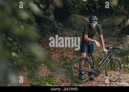 Porträt eines jungen Mannes, der mit einem schwarzen Fahrrad im grünen Stadtpark steht. Männlicher Athlet in Sportkleidung, Schutzhelm und Brille. Aktiv h Stockfoto