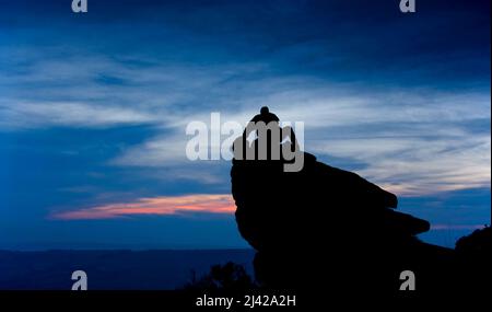 Nicht identifizierter Mann, der auf einem Felsvorsprung in Silhouette mit tiefblauem und rosafarbenem Abendhimmel hinter sich sitzt Stockfoto