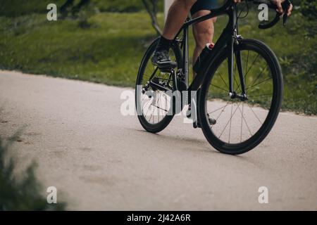 Nahaufnahme eines männlichen Athleten mit muskulösen Beinen, der auf einem schwarzen Fahrrad im grünen Park unterwegs ist. Mann in Sportswear und Sneakers, der morgens Zeit für das Training auf Fresh verbringt Stockfoto