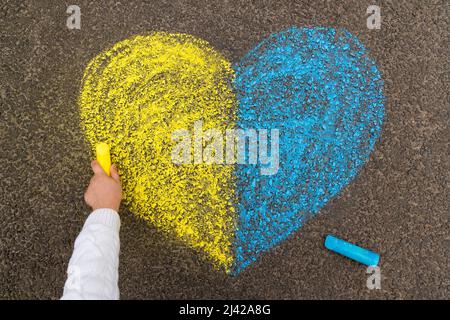 Die Hand eines Kindes zeichnet ein Herz mit Buntstiften auf dem Bürgersteig mit den patriotischen nationalen ukrainischen Farben Gelb und Blau. Beendet den Krieg in der Ukraine. Hochwertige Fotos Stockfoto