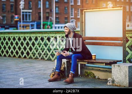 Das Warten auf den Bus muss keine Bohrung sein. Aufnahme eines jungen Mannes, der ein digitales Tablet benutzt, während er an einer Bushaltestelle in der Stadt wartet. Stockfoto