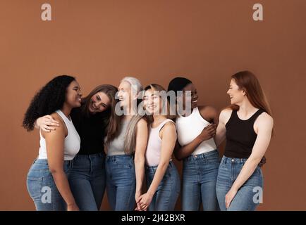 Sechs lachende Frauen verschiedener Körpertypen und Ethnien im Studio. Gruppenporträt lächelnder Frauen, die vor braunem Hintergrund zusammenstehen. Stockfoto