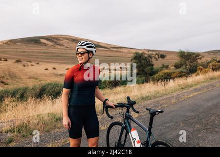 Die Sportlerin trägt einen Fahrradhelm und eine Sonnenbrille und steht mit einem Fahrrad auf einer leeren Straße Stockfoto