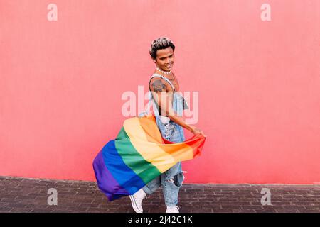 Lächelnder, gutaussehender Kerl, der mit LGBT-Flagge gegen die rosa Wand im Freien läuft Stockfoto