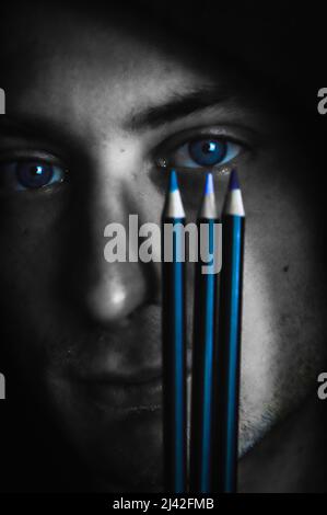 Passende Farben - Schwarz-Weiß-Porträt eines jungen blauäugigen Mannes, der drei Buntstifte in der Nähe seines Auges hält, um den richtigen Farbton zu finden Stockfoto