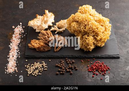 Rohe Sparassis (Blumenkohlpilz), Hericium, Clavaria (Korallenpilz) auf schwarzem Betongrund mit Salz und Gewürzen. Seitenansicht. Stockfoto