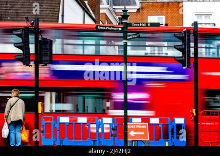 Epsom Surrey London, Großbritannien, 11 2022. April, eine Frau, die auf die Cross Road wartet, während der rote Doppeldeckerbus an der Ampel vorbeifährt Stockfoto