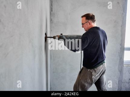 Seitenansicht eines erwachsenen Baumeisters mit einer Brille, der bei der Überholung des Wohnraums mithilfe eines Perforators ein Loch in der grauen Wand im Raum macht. Rekonstruktionsarbeiten in der Wohnung. Stockfoto