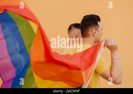 Junge schwule Paar hält Regenbogenfahne, während sie über der gelben Wand steht Stockfoto