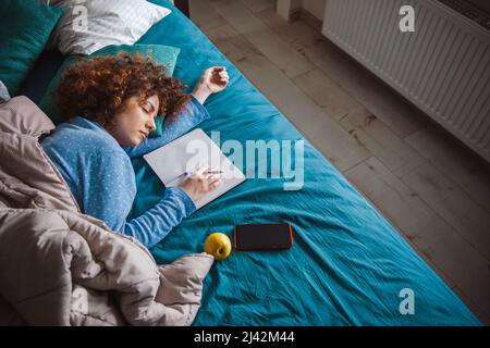 Lockiges Studentenmädchen, das mit Lehrbüchern auf dem Bett liegt und zu Hause glücklich schläft. Konzept zur Schlafenszeit. Komfortkonzept. Stockfoto