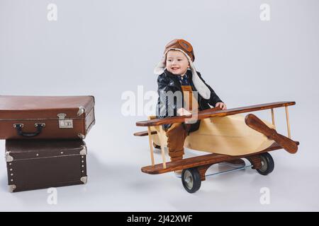 Ein kleiner Junge sitzt auf einem hölzernen Flugzeug. Umweltfreundliches Kinderspielzeug. Junge in Lederjacke und Hut Stockfoto