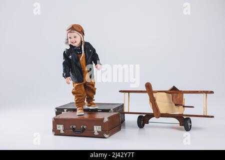 Ein glücklicher Junge in einem Helm und einer Pilotenjacke steht in der Nähe eines Holzflugzeugs. Porträt eines Pilotin-Kindes, eines Kindes in einer Lederjacke. Holzspielzeug. Öko-Plan Stockfoto