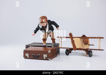 Ein glücklicher Junge in einem Helm und einer Pilotenjacke steht in der Nähe eines Holzflugzeugs. Porträt eines Pilotin-Kindes, eines Kindes in einer Lederjacke. Holzspielzeug. Öko-Plan Stockfoto