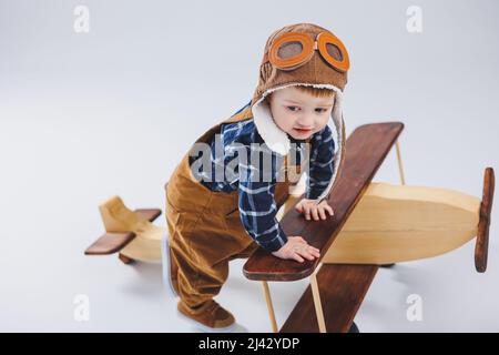 Kleiner Junge 3 Jahre alt spielt mit einem hölzernen Flugzeug auf weißem Hintergrund. In Overalls und einem Pilotenhut. Öko Spielzeug für Kinder aus Naturholz. woode Stockfoto