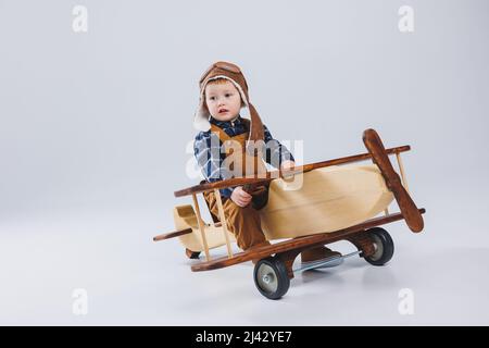 Kleiner Junge 3 Jahre alt spielt mit einem hölzernen Flugzeug auf weißem Hintergrund. In Overalls und einem Pilotenhut. Öko Spielzeug für Kinder aus Naturholz. woode Stockfoto