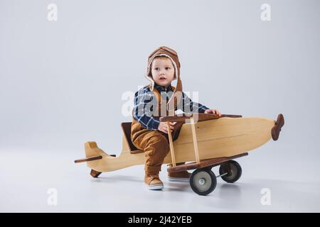 Kleiner Junge 3 Jahre alt spielt mit einem hölzernen Flugzeug auf weißem Hintergrund. In Overalls und einem Pilotenhut. Öko Spielzeug für Kinder aus Naturholz. woode Stockfoto