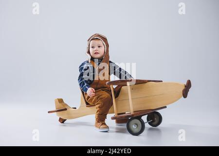 Kleiner Junge 3 Jahre alt spielt mit einem hölzernen Flugzeug auf weißem Hintergrund. In Overalls und einem Pilotenhut. Öko Spielzeug für Kinder aus Naturholz. woode Stockfoto