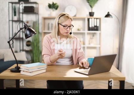 Porträt von schönen selbstbewusst mit blonden Haaren und auf Kopfhörern clevere Frau in legerer Kleidung, sie sitzt am Tisch vor dem Computer und überprüfen Informationen in Präsentation im Internet Stockfoto