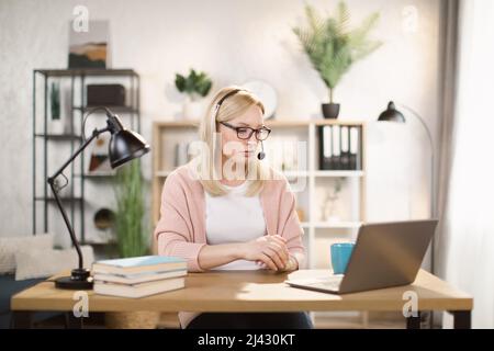 Porträt von schönen selbstbewusst mit blonden Haaren und auf Kopfhörern clevere Frau in legerer Kleidung, sie sitzt am Tisch vor dem Computer und überprüfen Informationen in Präsentation im Internet Stockfoto