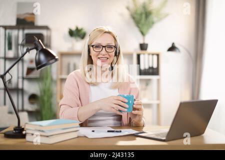 Eine ziemlich reife kaukasische Frau im Headset trinkt heißen Tee oder Kaffee und lächelt, während sie mit einem Laptop online in einem hellen, modernen Zimmer zu Hause arbeitet. Fernarbeit und Bildung während der Pandemie Stockfoto