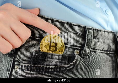 Eine Frauenhand steckt eine goldene Bitcoin in eine Jeanstasche, Nahaufnahme Stockfoto