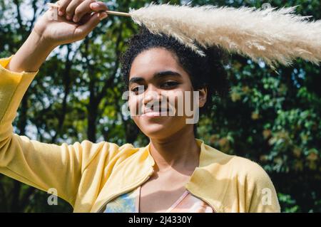 Portrait schönes junges schwarzes Mädchen mit afro lächelnd sehr glücklich in der Natur, das mit der Hand ein Pampagras über sie hält und die Kamera anschaut. Stockfoto