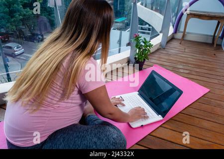 Rückansicht einer Schwangeren, die zu Hause auf einer rosa Yogamatte sitzt und auf einer Laptop-Tastatur tippt, Yoga praktiziert und meditiert Stockfoto
