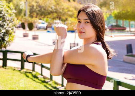 Eine junge kaukasische Athletin dehnt ihre Schultermuskeln in einem Park. Das Mädchen trägt ein Activity-Armband. Stockfoto