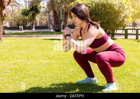 Eine junge kaukasische Athletin hockt auf dem Gras in einem Park. Das Mädchen trägt ein Activity-Armband. Stockfoto