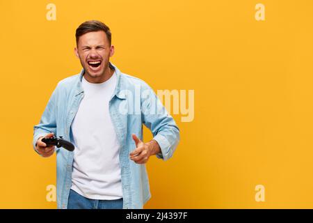 Irritisierte böse gebräunte gutaussehende Mann in blauem Basis-T-Shirt wütend über Verlust halten Joystick Gamepad posiert isoliert auf orange gelben Studio-Hintergrund Stockfoto