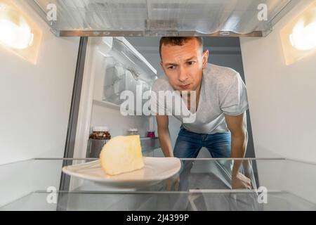 Der Mann sieht auf einen Teller Käse in einem leeren Kühlschrank. Konzept der Lieferservice, Hunger, Diäten. Foto aus dem Kühlschrank Stockfoto