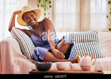 Es ist eine Haltung, die ich umarmte. Porträt einer jungen, schönen Frau, die einen Sonnenhut trägt, während sie sich zu Hause auf dem Sofa entspannt. Stockfoto